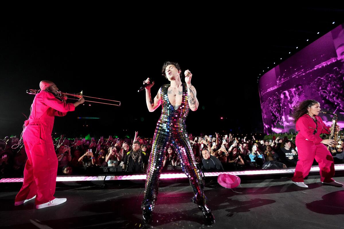 A male singer in a shiny jumpsuit performs onstage with two female horn players in pink coveralls.