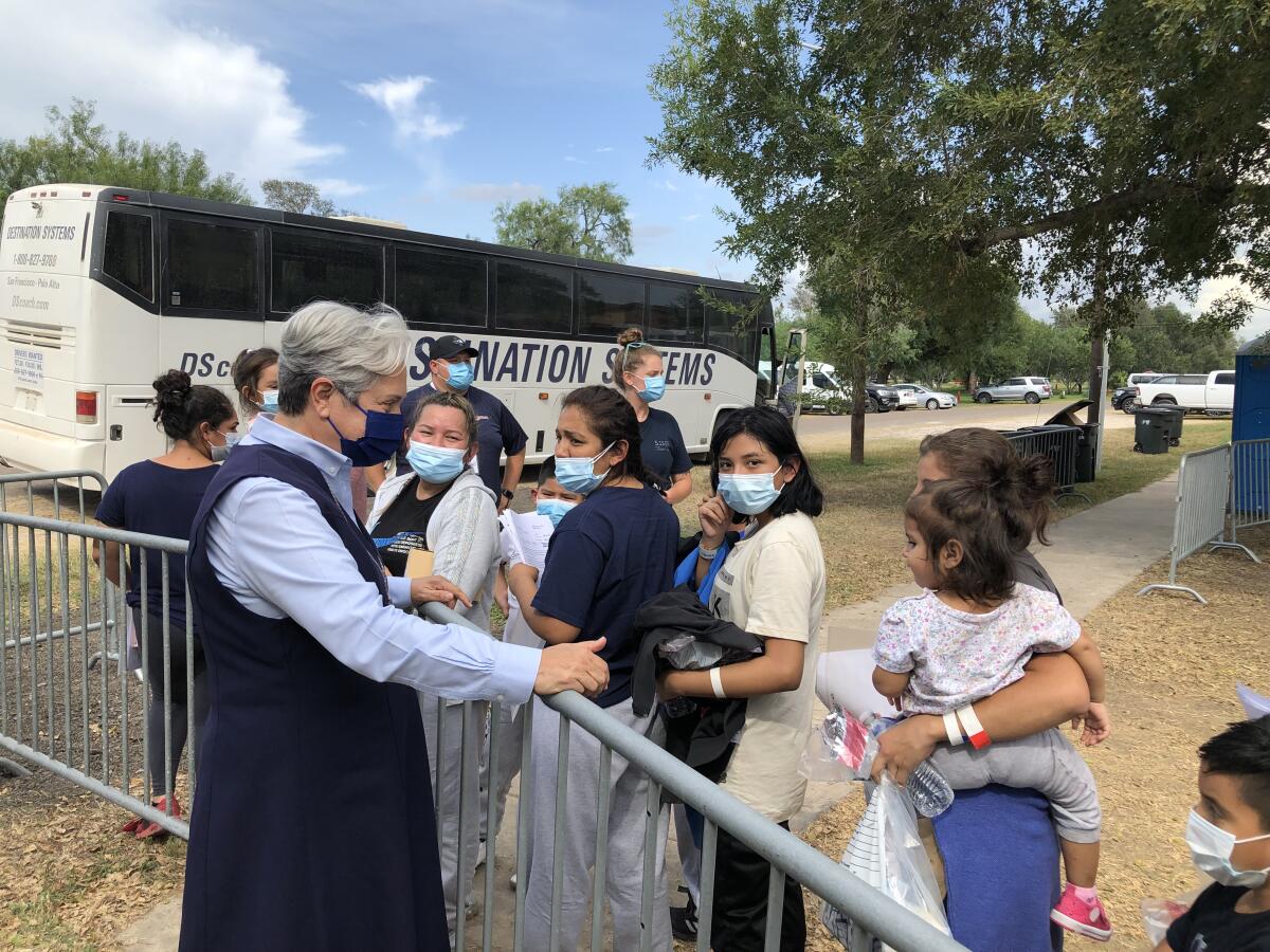 A woman speaks to people standing in line with a bus in the background.