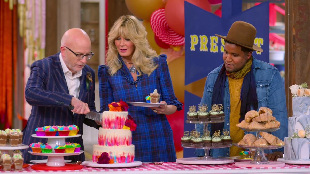 A woman flanked by two men holds a plate as they all look at cakes and cupcakes on a table.