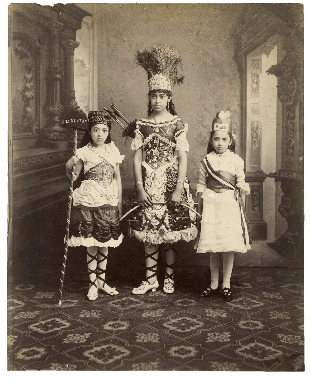 A sepia image shows three little girls in fantastical outfits, one with a headdress, in a carpeted foyer