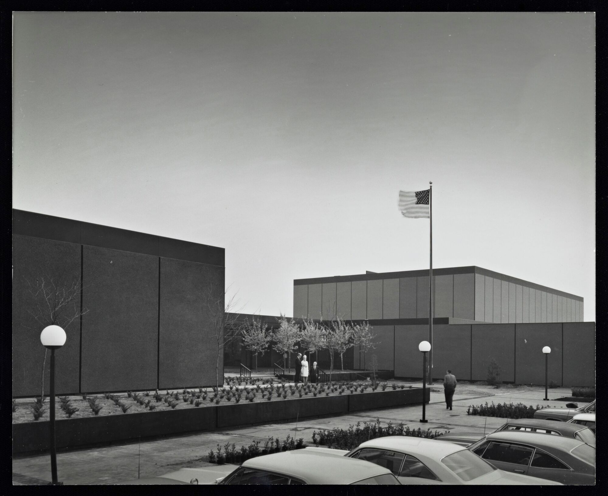 A vintage black and white image shows two blocky buildings separated by a breezeway, over which fly a U.S. flag