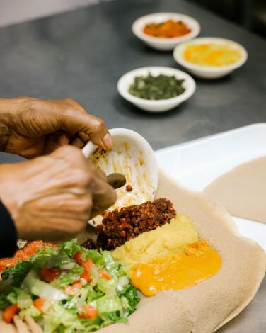 Hands prepare a sampler of Ethiopian foods on a plate covered with injera bread