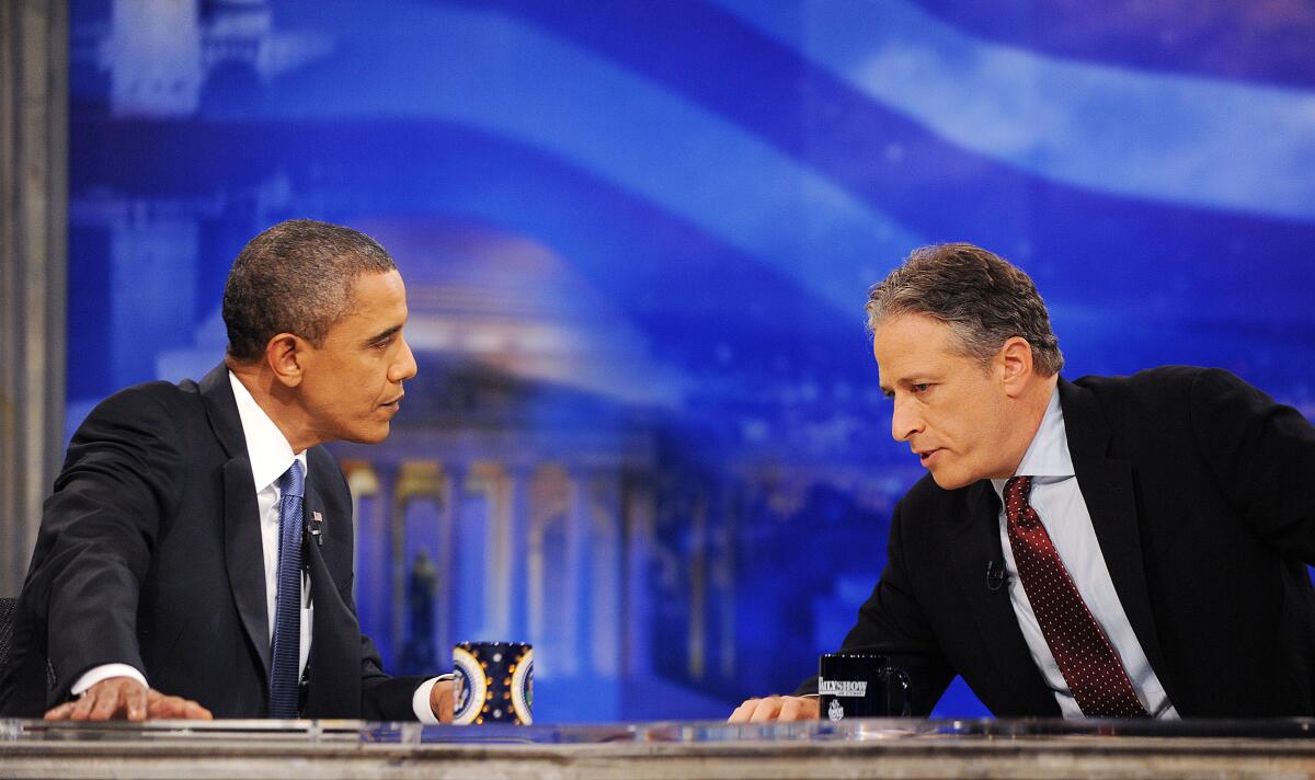 Jon Stewart leans over his desk to talk to President Obama