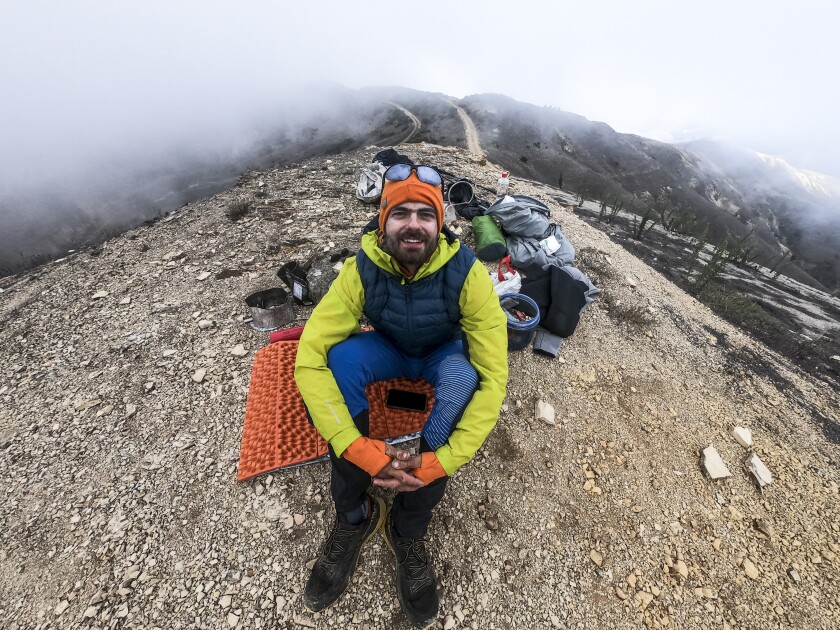 A young man sits on a hilltop surrounded by hiking and camping equipment.