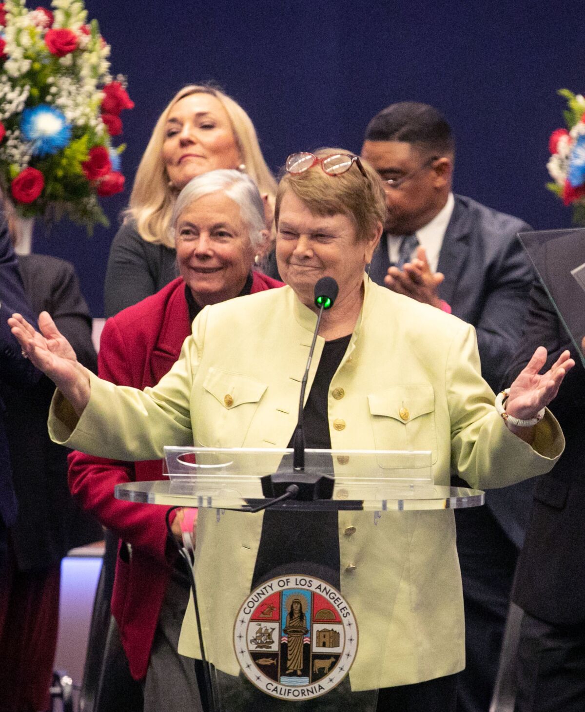 Sheila Kuehl smiles at a lectern and raises her hands while colleagues stand behind her.