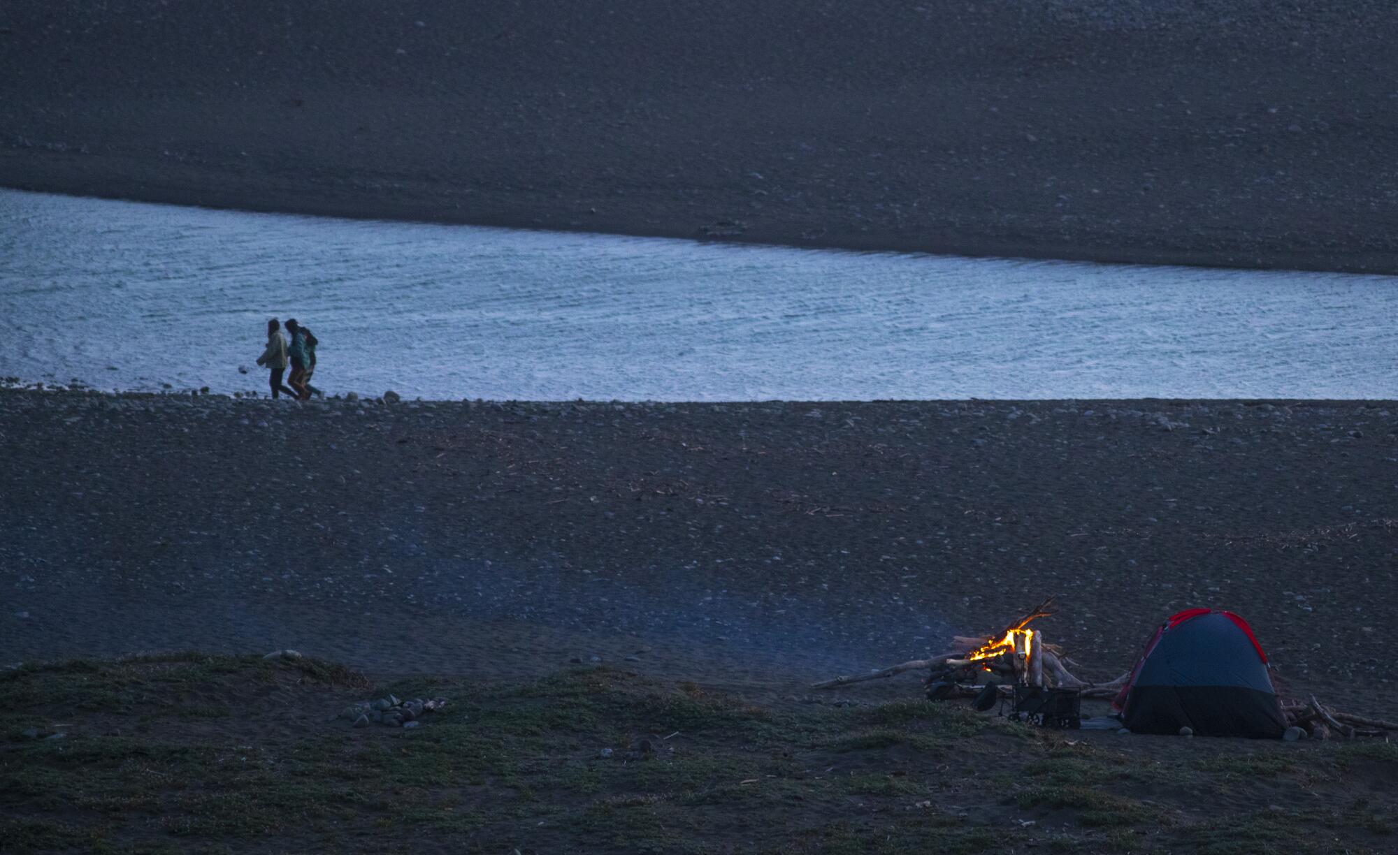 People are seen on a flat, wide, rocky beach at dusk; in the foreground a campfire burns next to a small tent.