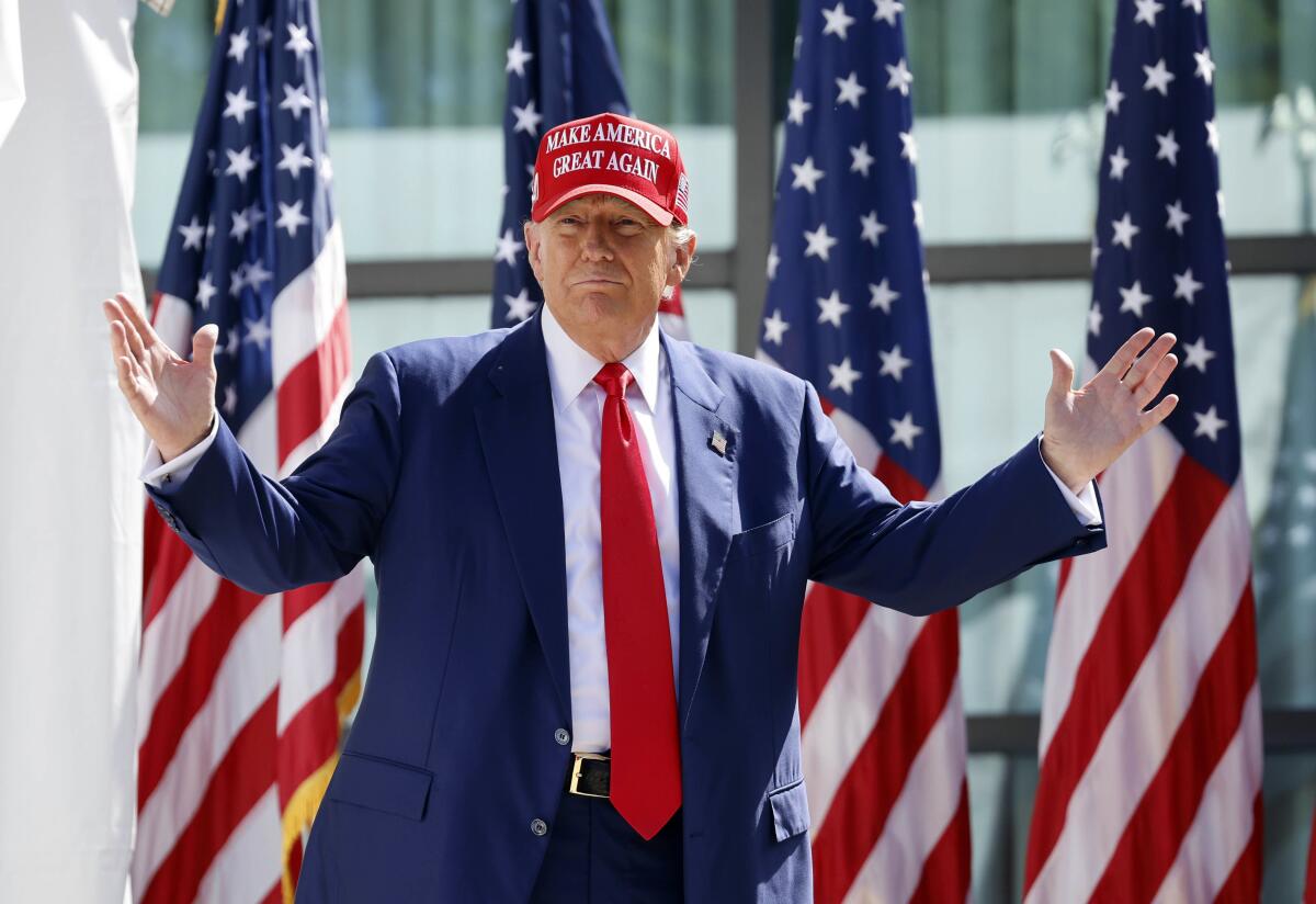 Donald Trump, wearing a suit and a red MAGA hat, raises his arms in front of American flags. 