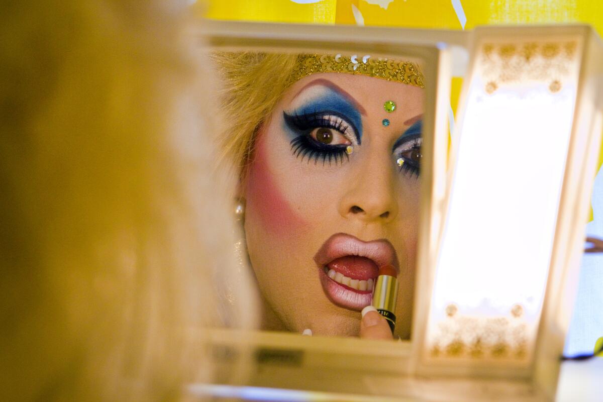 A drag queen applies lipstick while looking at her reflection in a mirror