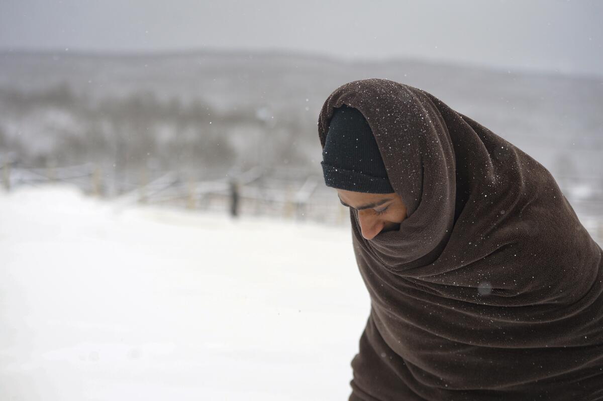 A man in beanie and wrapped in a blanket walks through a snowy field.
