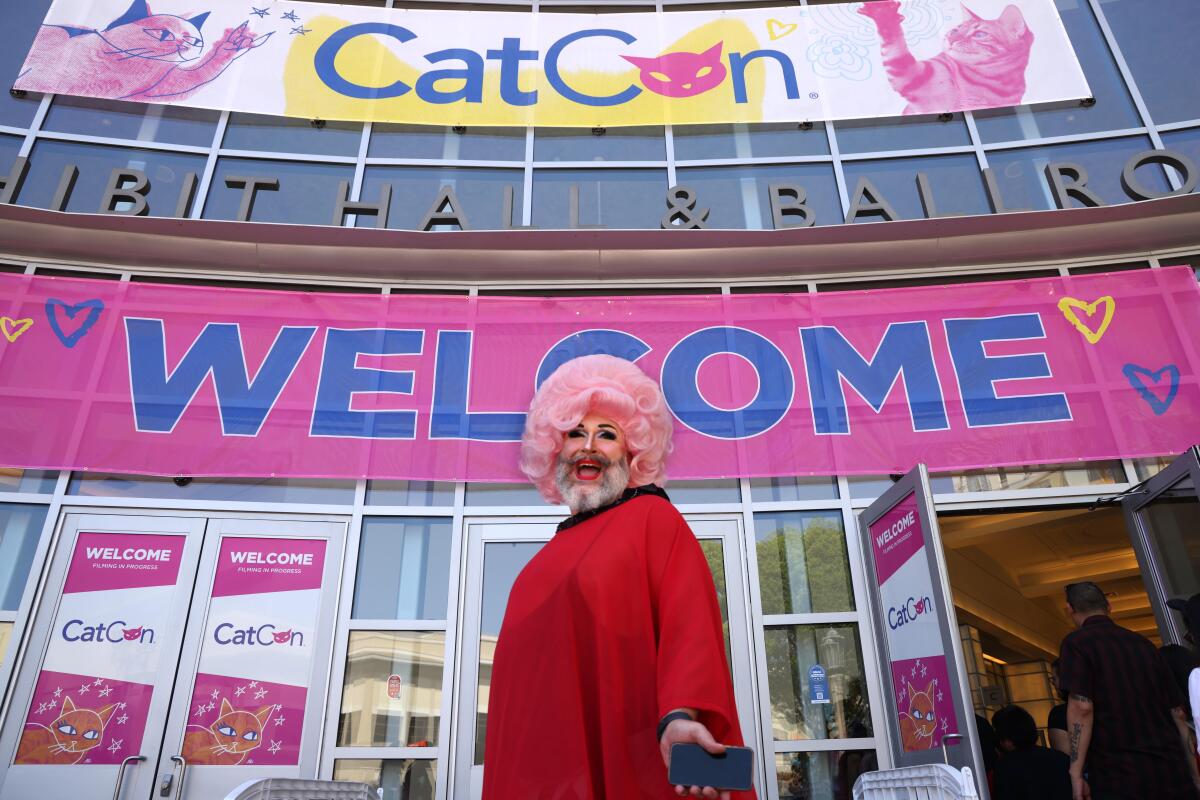 A person in a red robe and a wavy pink wig, standing in front of a "Welcome" Sign up for CatCon.