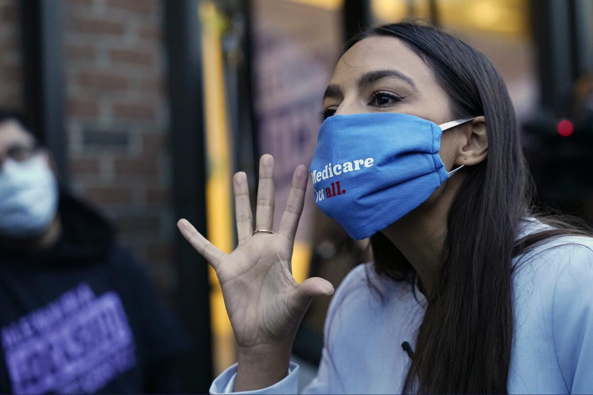 Rep. Alexandria Ocasio-Cortez gestures with her hand while wearing a Medicare for all mask.