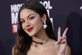 A woman with long brown hair wearing red lipstick and dangly earrings while making a peace sign with her left hand