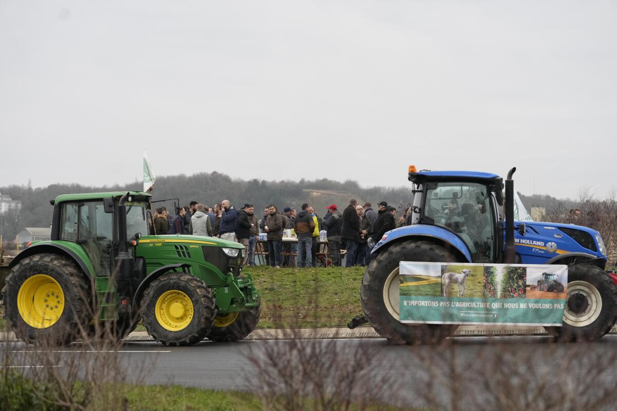 Two tractors stopped on a paved road under a gray sky as a small crowd stands on the turf in the background