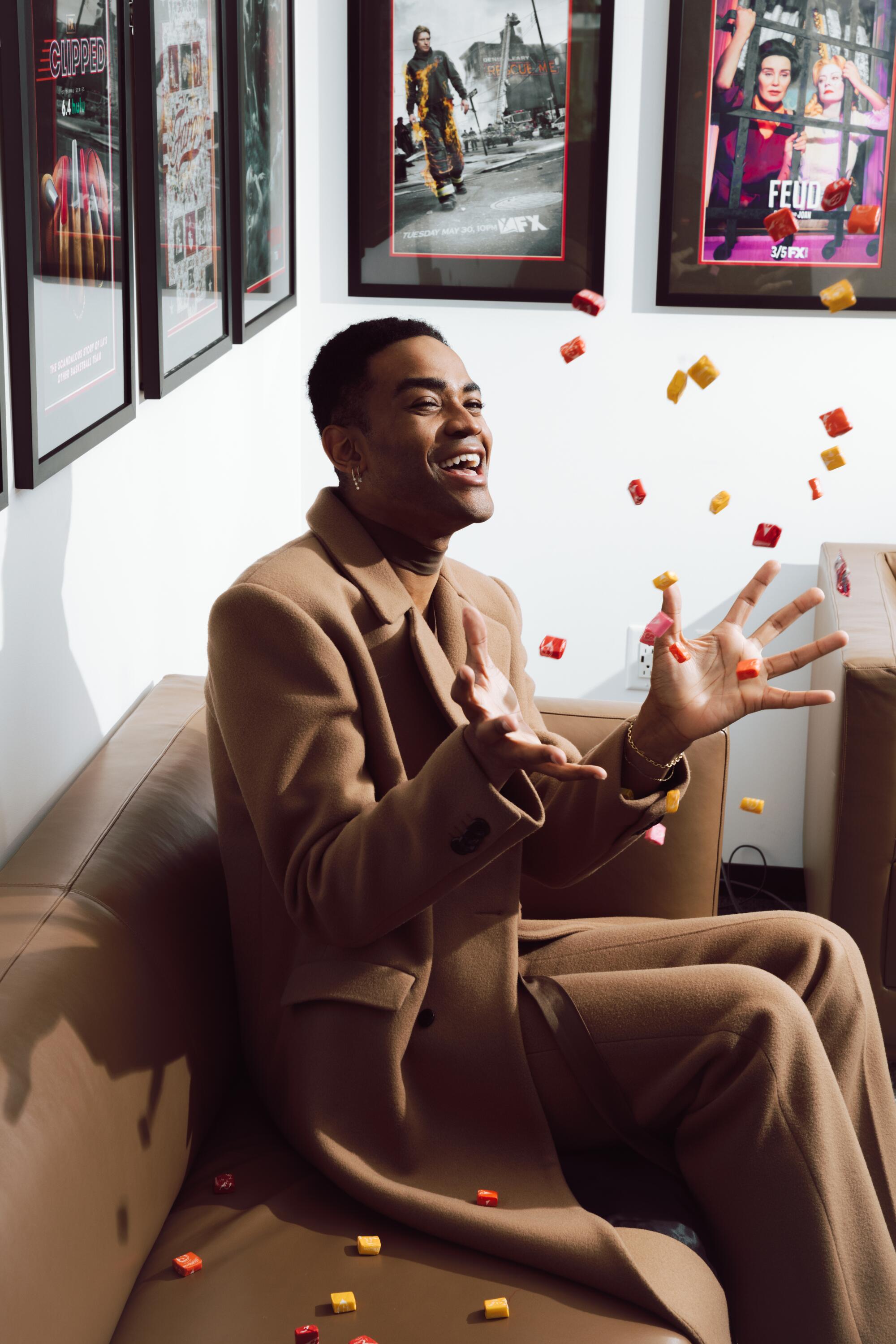 A man in a tan coat and slacks sitting on a couch throwing many Starburst candies in the air.