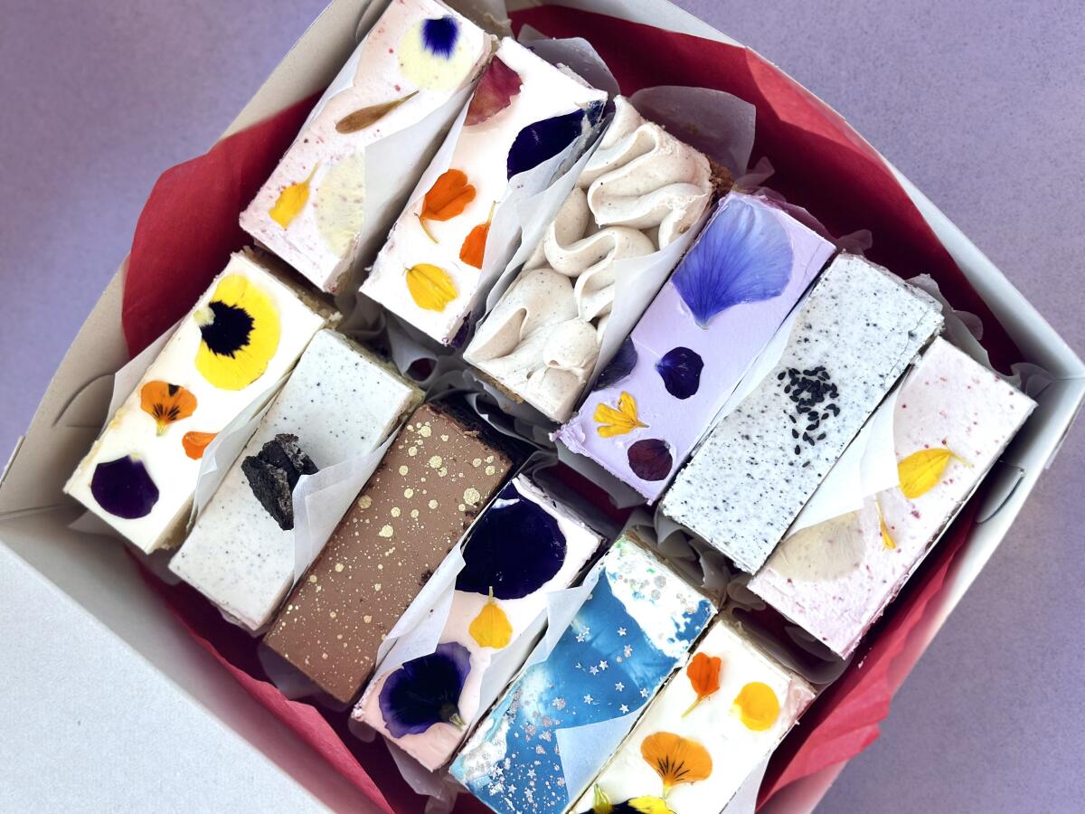 A dozen different rectangular cake bars, decorated with frosting, flower petals and sprinkles, in a box