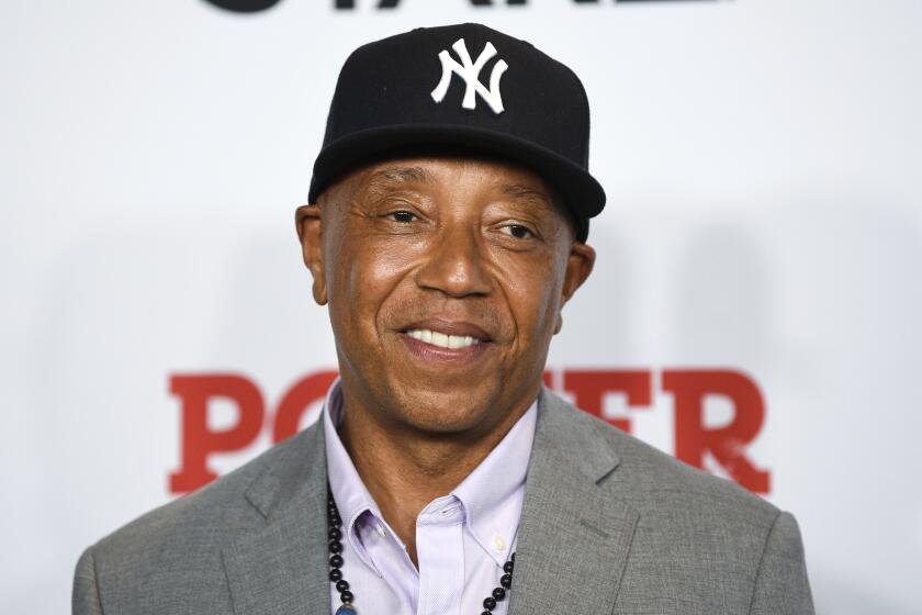 A Black man in a black New York Yankees hat smiling and wearing a gray blazer, white suit shirt and a beaded necklace