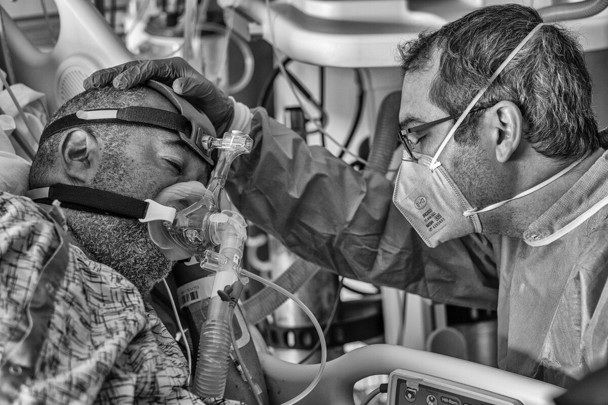 A man in a mask holds his hand on the head of a man in a hospital bed who is hooked up to a respirator.