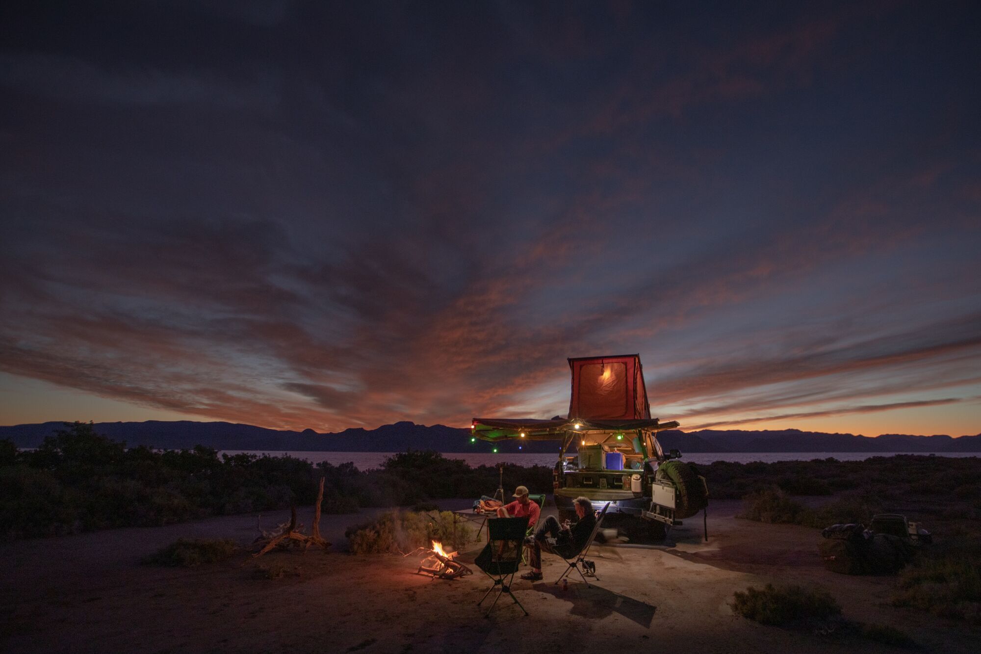 Two men sitting in chairs around a campfire at dusk, with their camper lit up with holiday lights and a bay behind them.