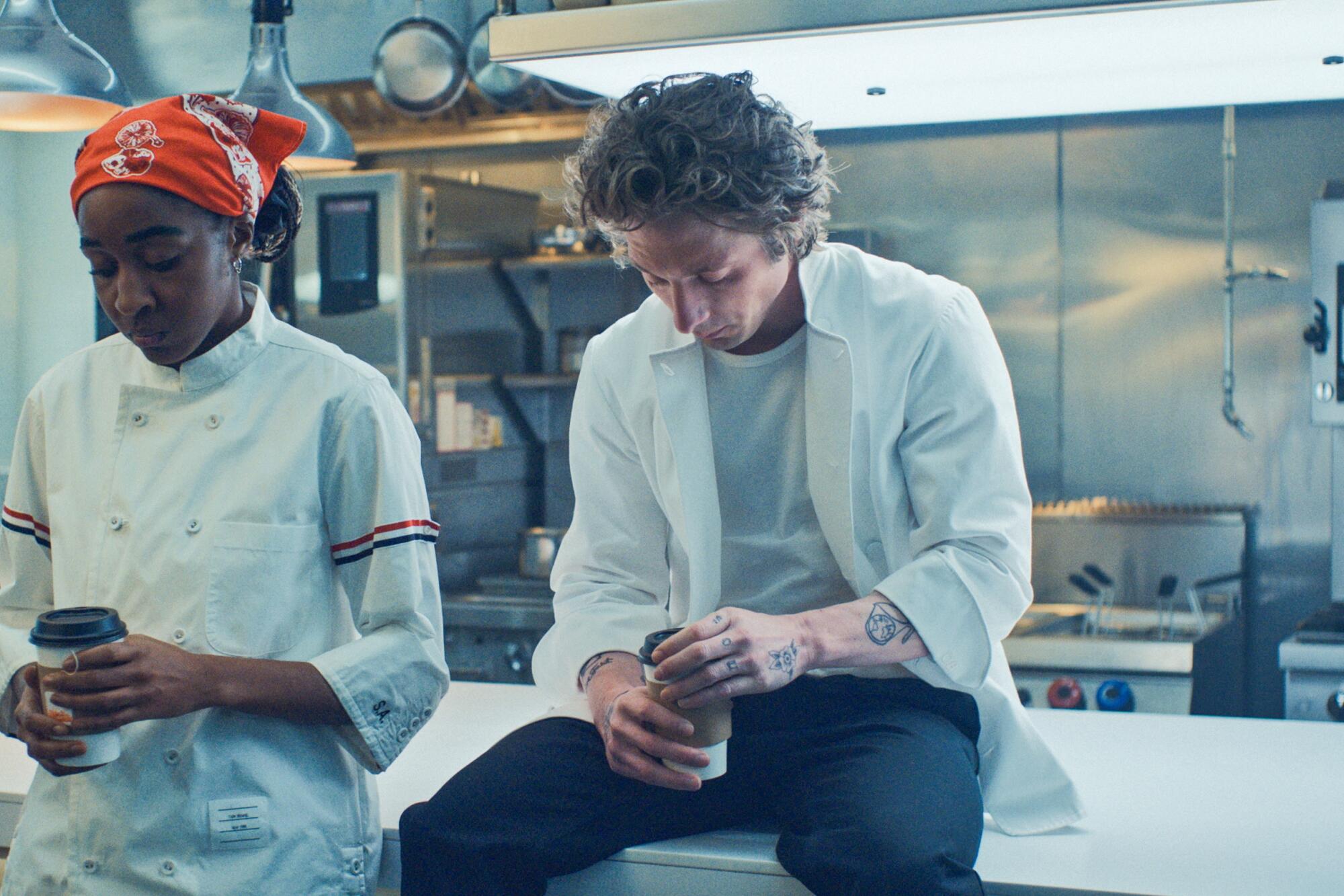 A man and woman in chef's jackets look down at their coffees while in an industrial kitchen in "The Bear."