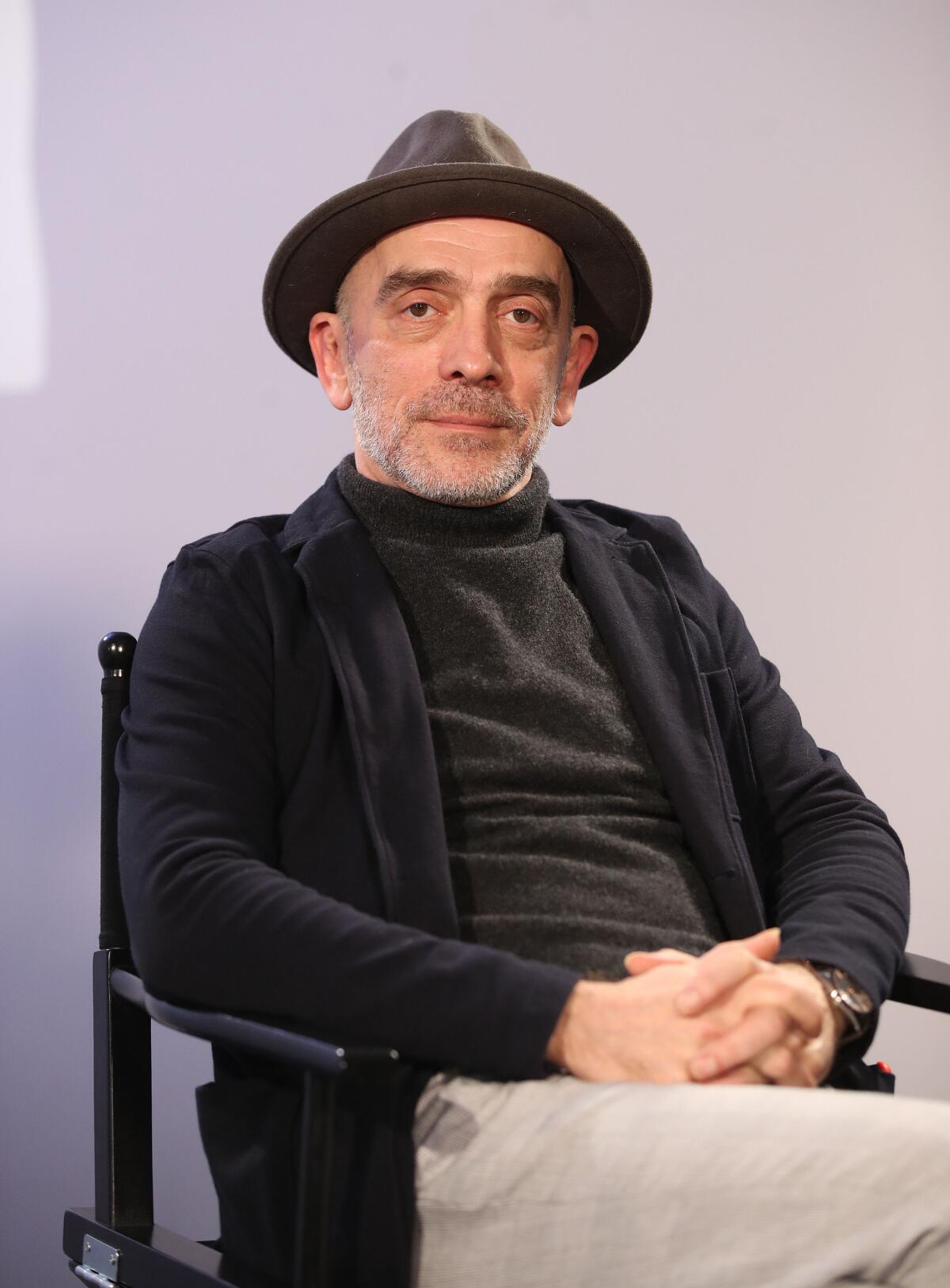 A man with short, gray facial hair in a wide-brimmed hat, a dark sweater and shirt sitting with his hands on his lap 