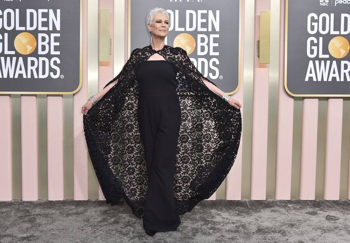 A woman holds her black lace cape out behind her with both hands while posing on a gray carpet