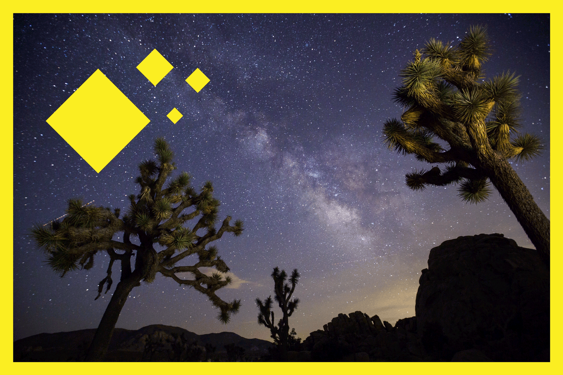 A view of the Milky Way arching over Joshua Trees and rocks at a park campground.