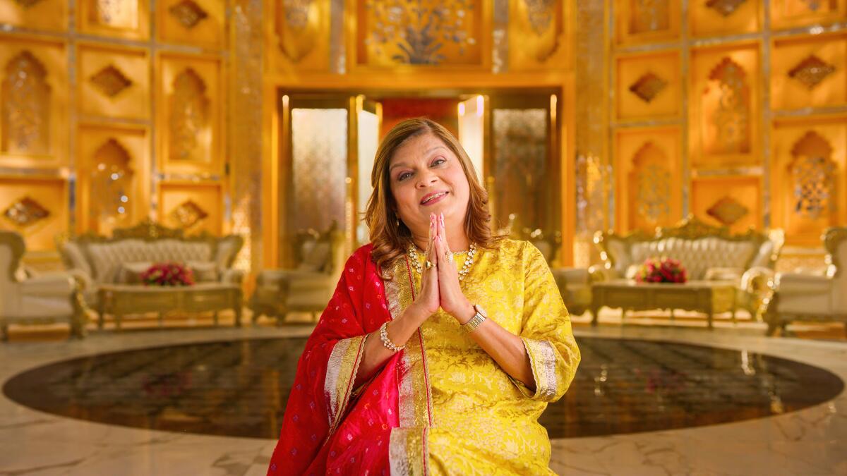 An Indian woman sits in an ornate gold room with her hands pressed together 