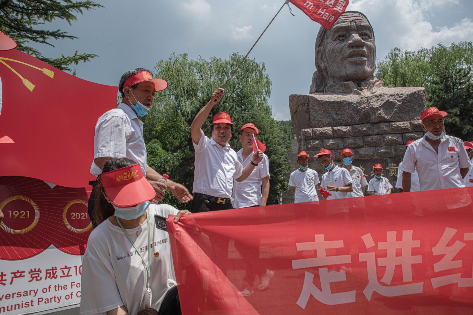 People in red visors and white shirts mill about near a large stone bust of a person  