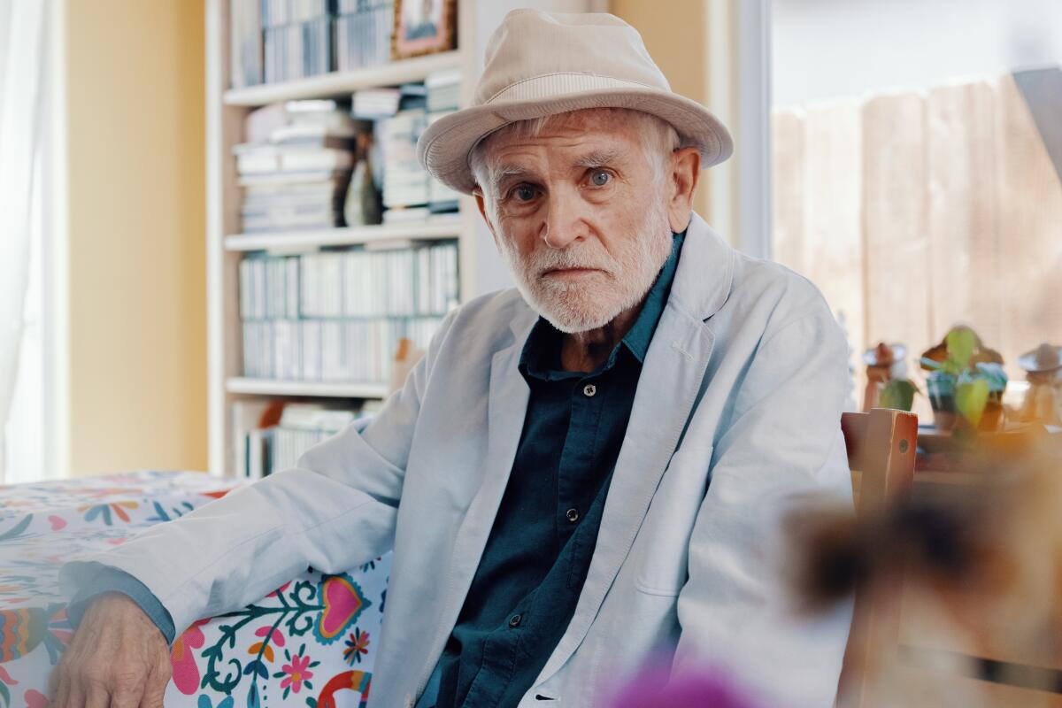 A white-bearded man in a hat and blazer sits on a sofa with a vibrant floral design.