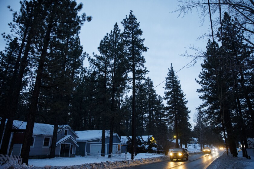 Cars with headlights on drive on a two-lane road between trees and snow-covered houses