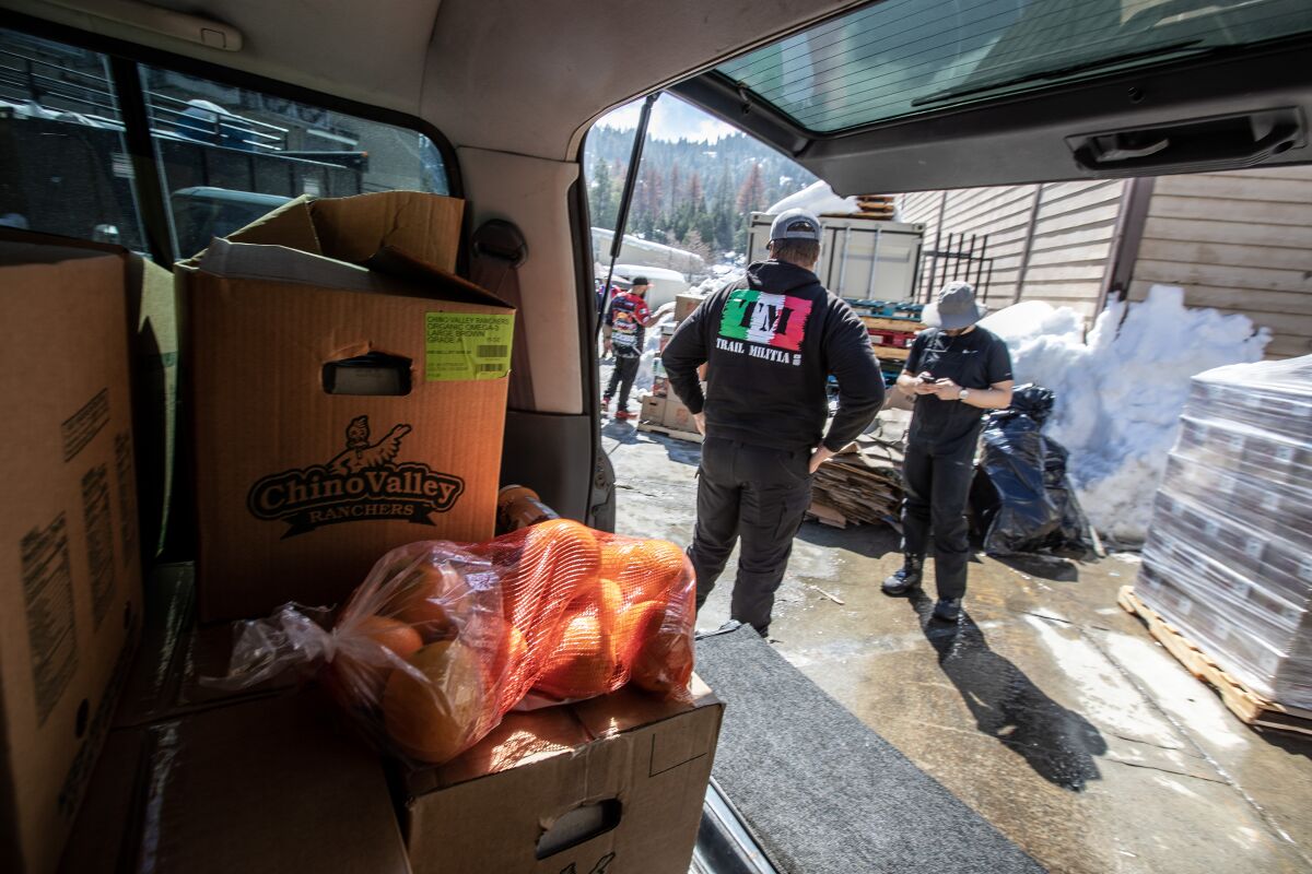 Boxes of groceries in the back of a vehicle and people stand outside near it.