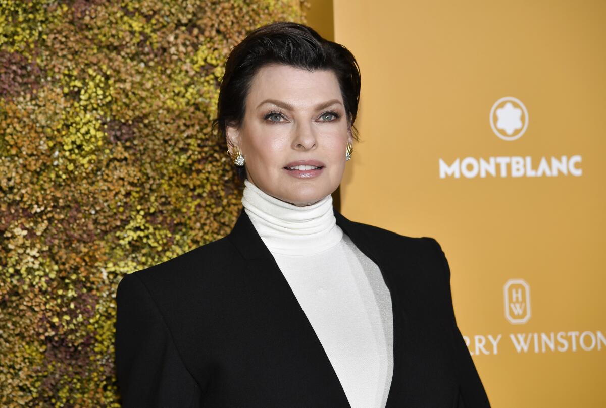 Linda Evangelista wears a white turtleneck and a black blazer, with short hair and sparkly earrings.