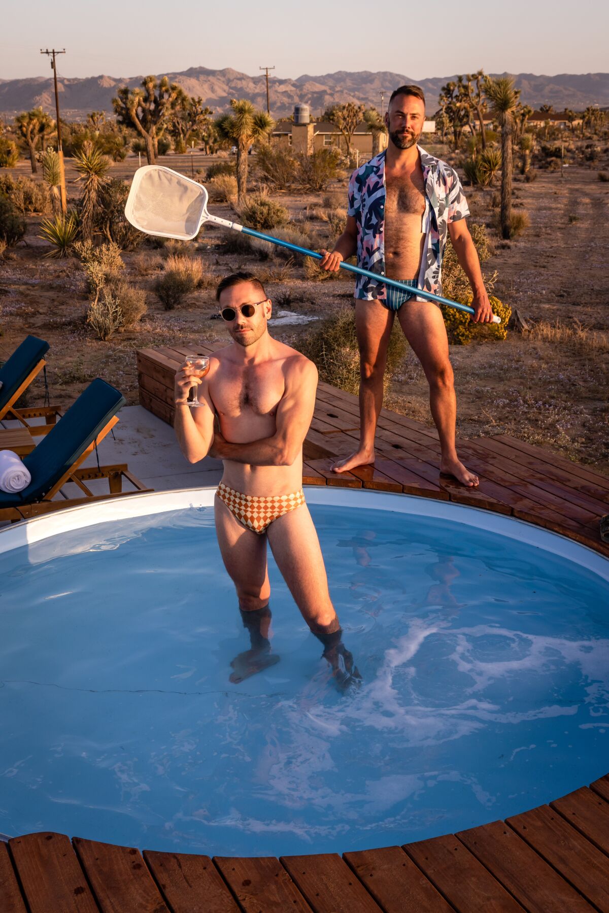 A man in a speedo in a round pool and a man behind him in a speedo holding a pool net 