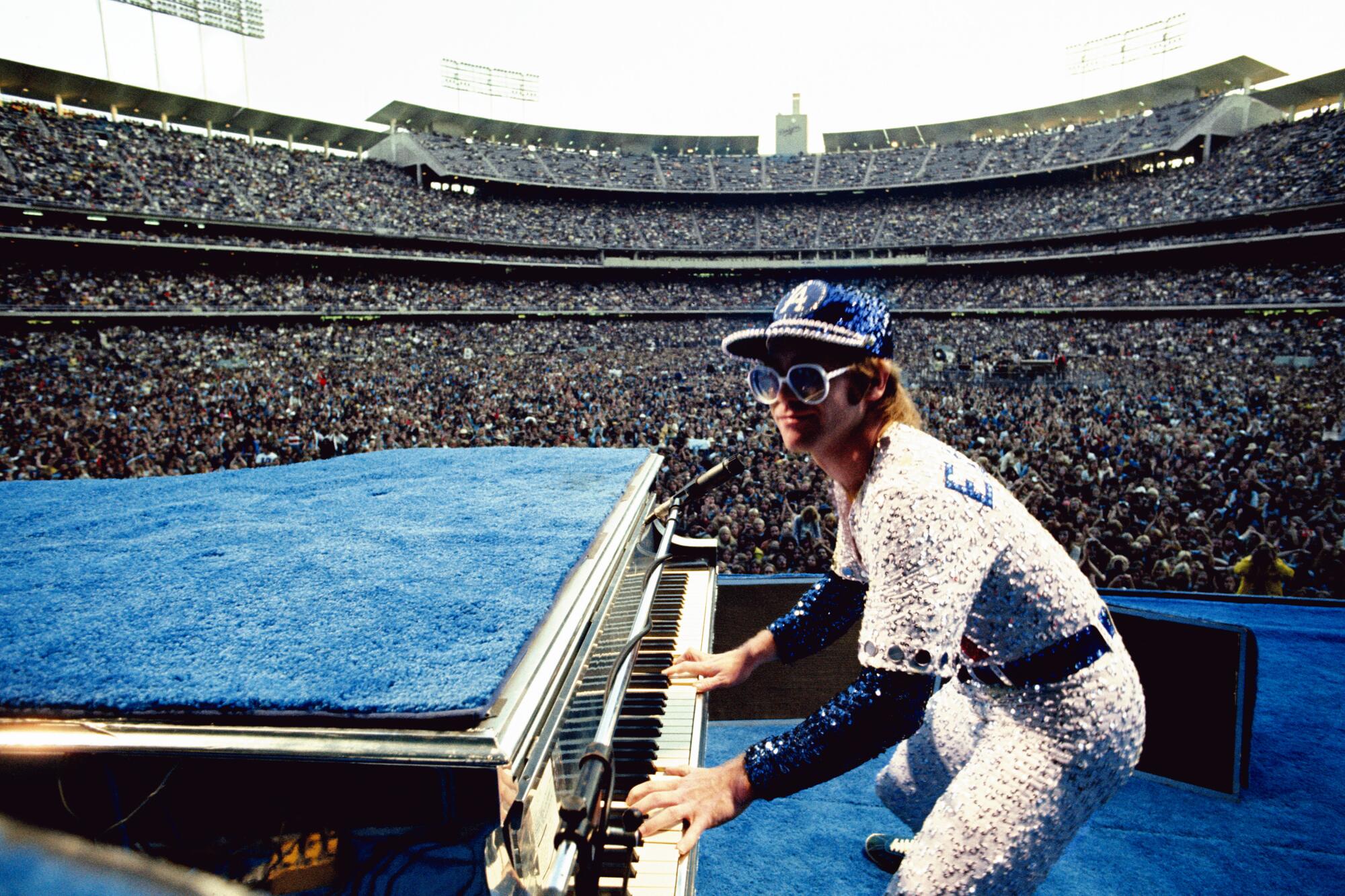 Elton John in a sequined Dodgers uniform plays the piano in front of a stadium full of fans.