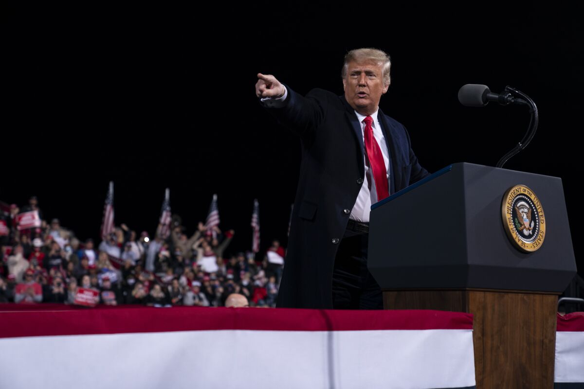 President Trump speaks at a lectern during a campaign rally with a crowd and U.S. flags behind him.