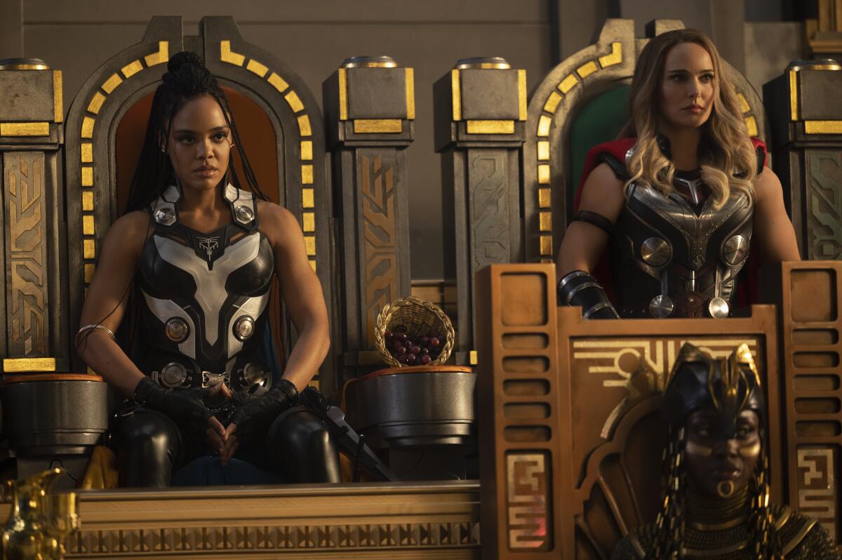 Two women in superhero costumes sit on gold-accented wooden thrones.