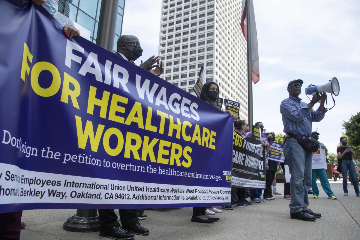 A man speaks into a megaphone as people behind him hold banners reading "Fair Wages for Healthcare Workers."