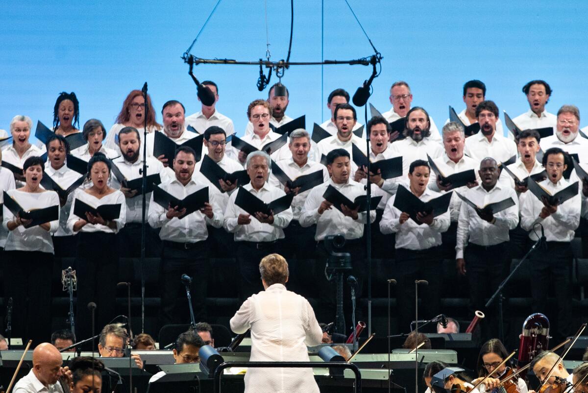 A woman is seen from behind conducting an orchestra and singers.