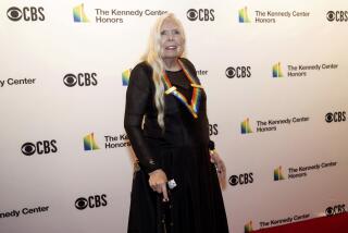 An older woman with long blond hair poses on a red carpet in a black dress