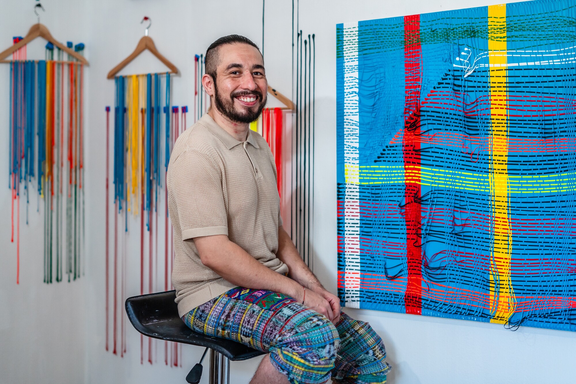 El artista Cristian García Olivo sentado frente a su pintura acrílica sobre una tabla de madera con escrito en ella "Artículos de primera necesidad" 