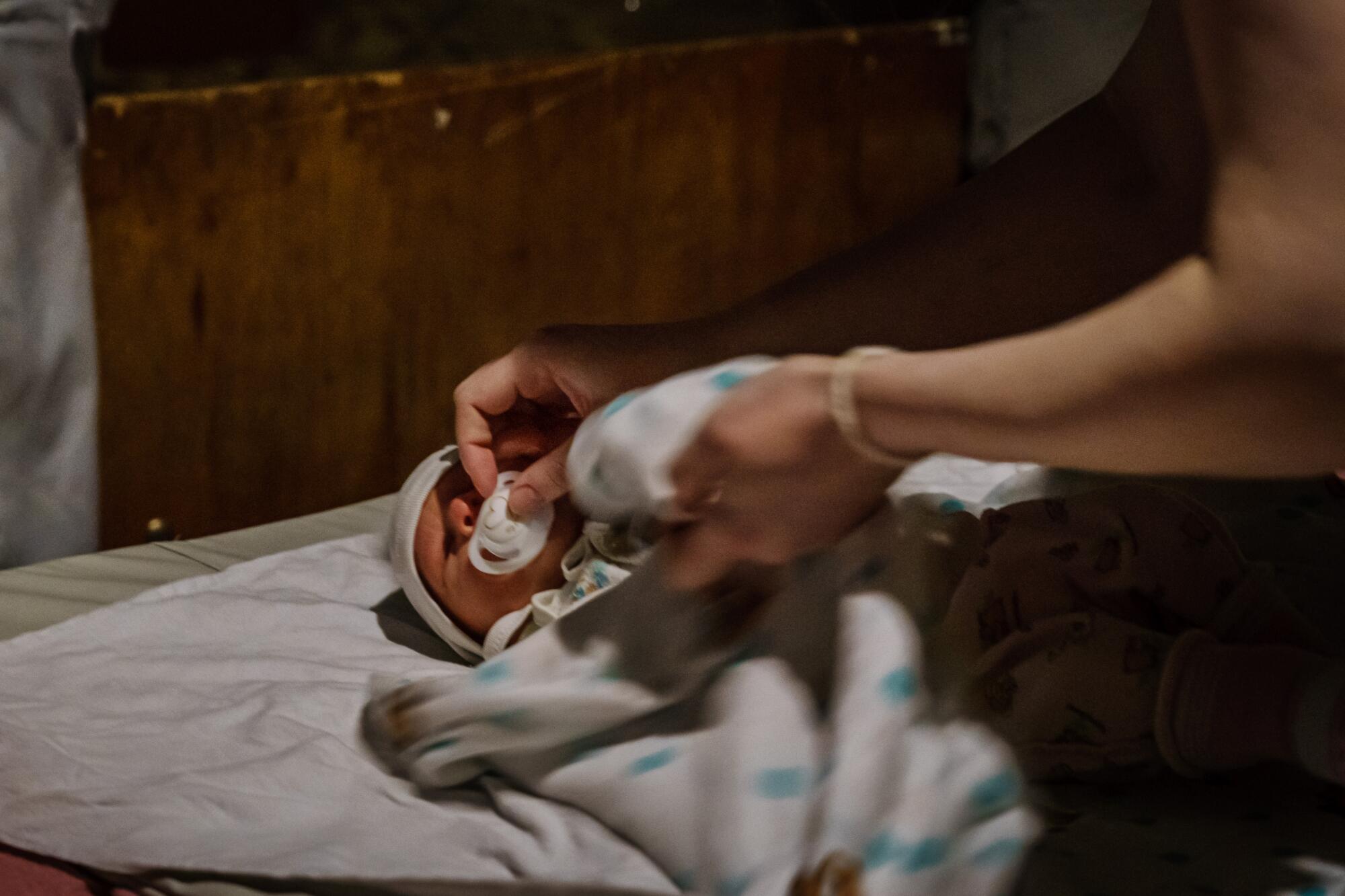 A baby in a white blanket lying down with a woman's hand placing a pacifier in the baby's mouth.