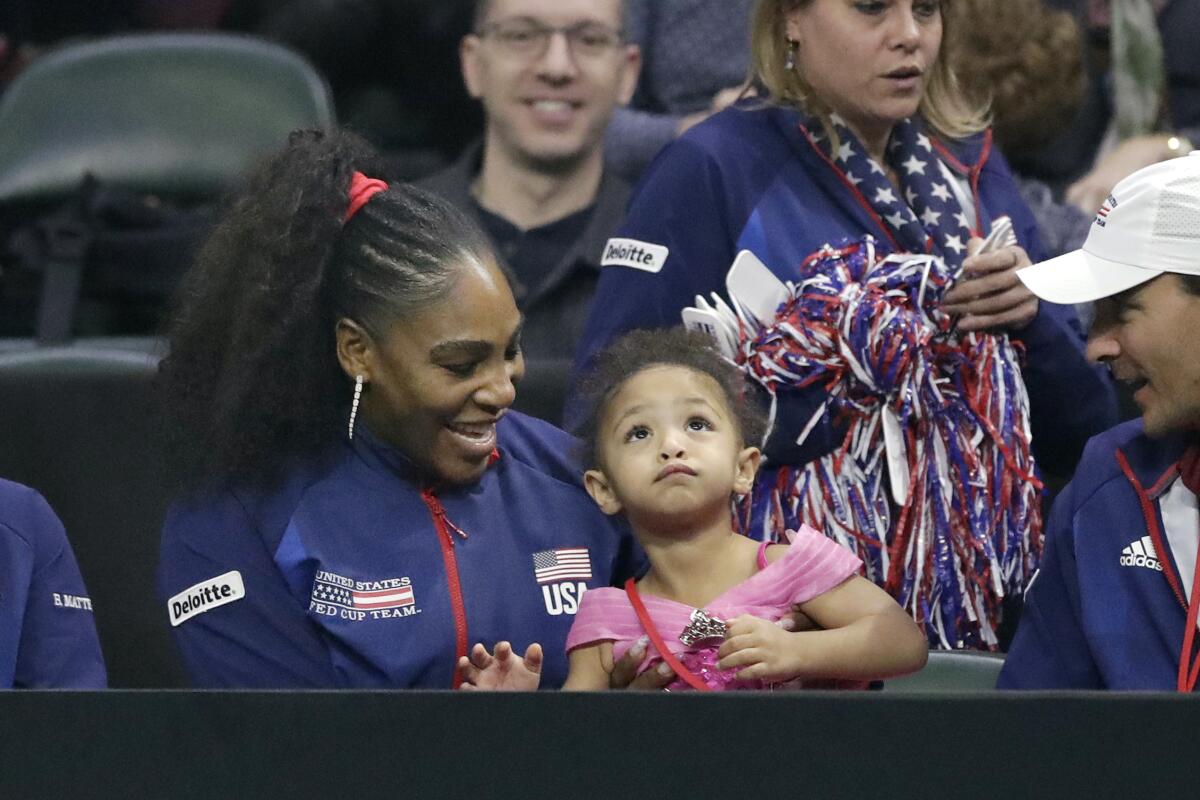 Serena Williams in a USA jacket sits among others with her young daughter, dressed in a pink princess dress, on her lap.