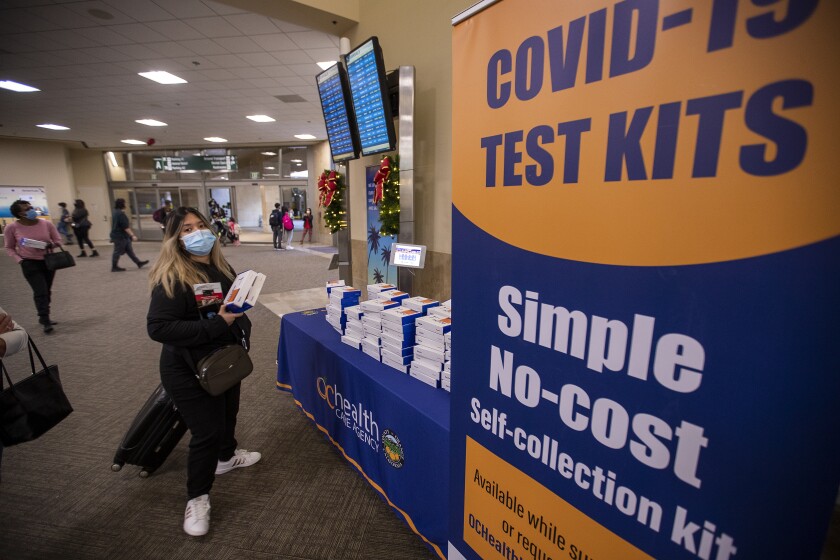 A woman in a mask stands holding boxes near a poster that says "COVID-19 test kits."