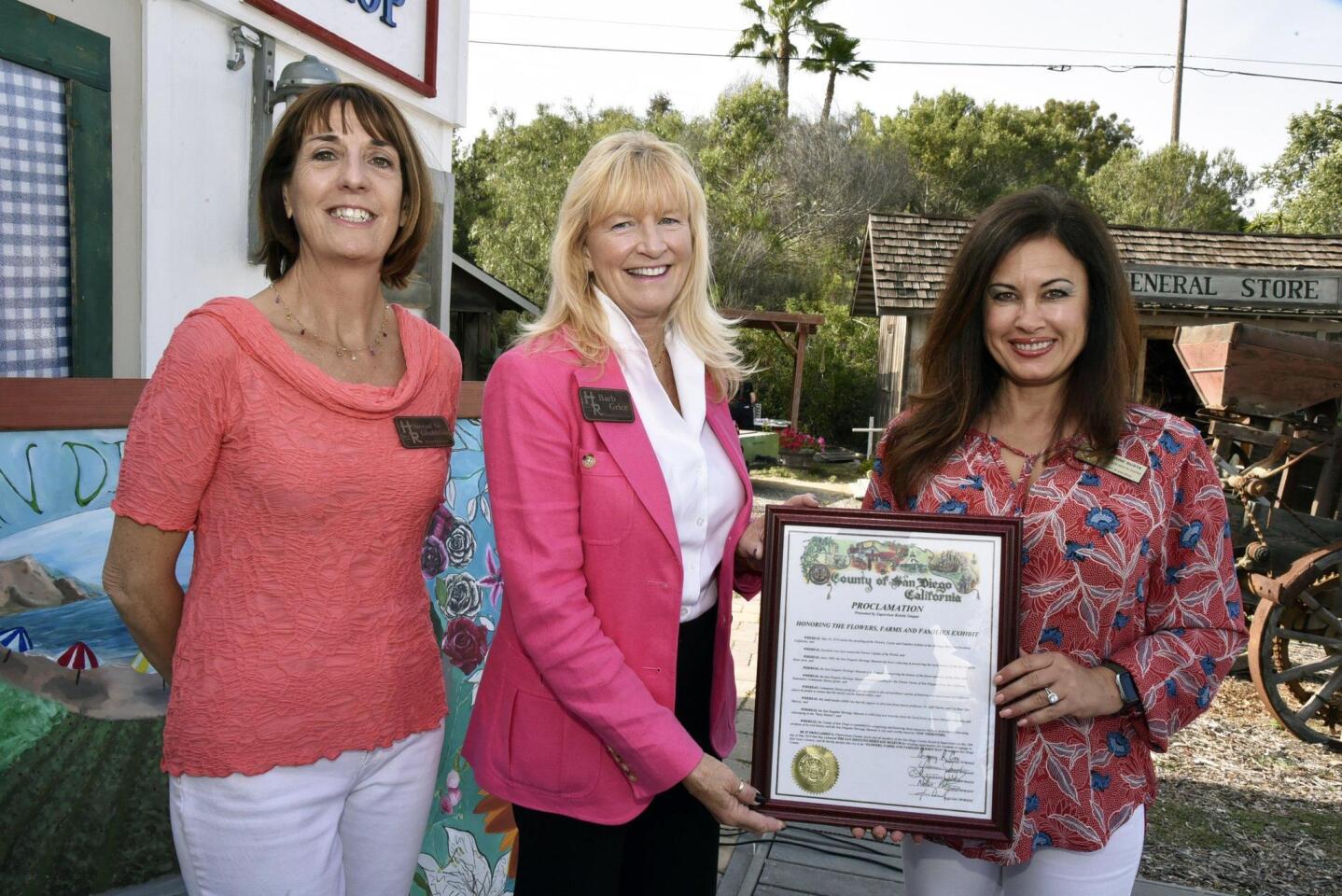 The San Dieguito Heritage Museum Board President Sinéad Ni Chabhláin and Executive Director Barb Grice receive a plaque honoring The Flowers, Farmers, and Families exhibit, presented by Corrine Busta (Policy Advisor representing Supervisor Kristin Gaspar)