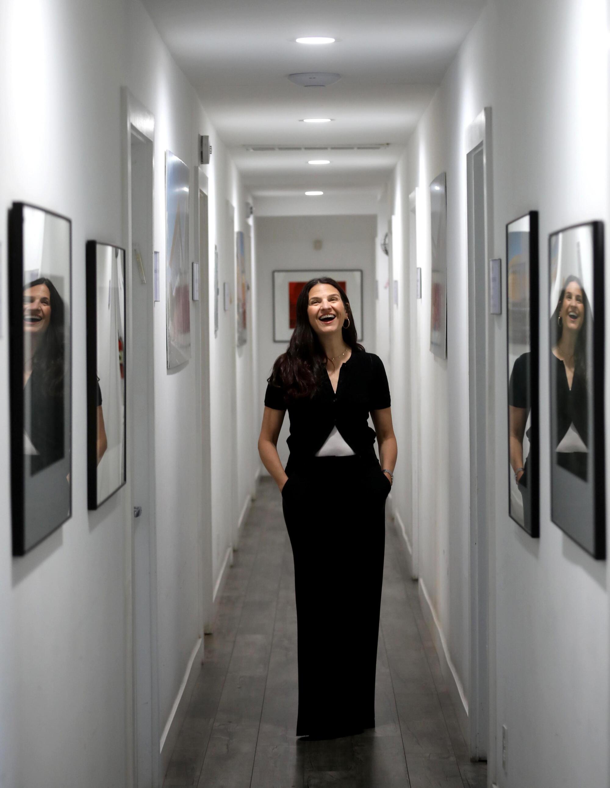 A woman stands in a corridor, her face reflected in the glass of large frames on the wall