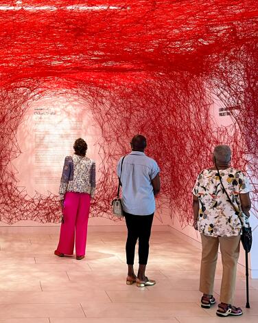 Three people stand inside an art exhibit at Hammer Museum. Red webbing covers the walls of the space.