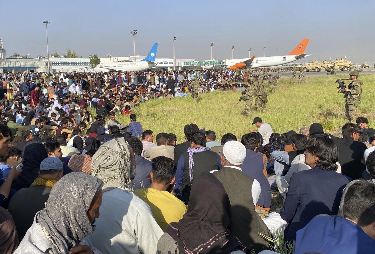 People sit on the ground near airplanes as troops stand and watch.