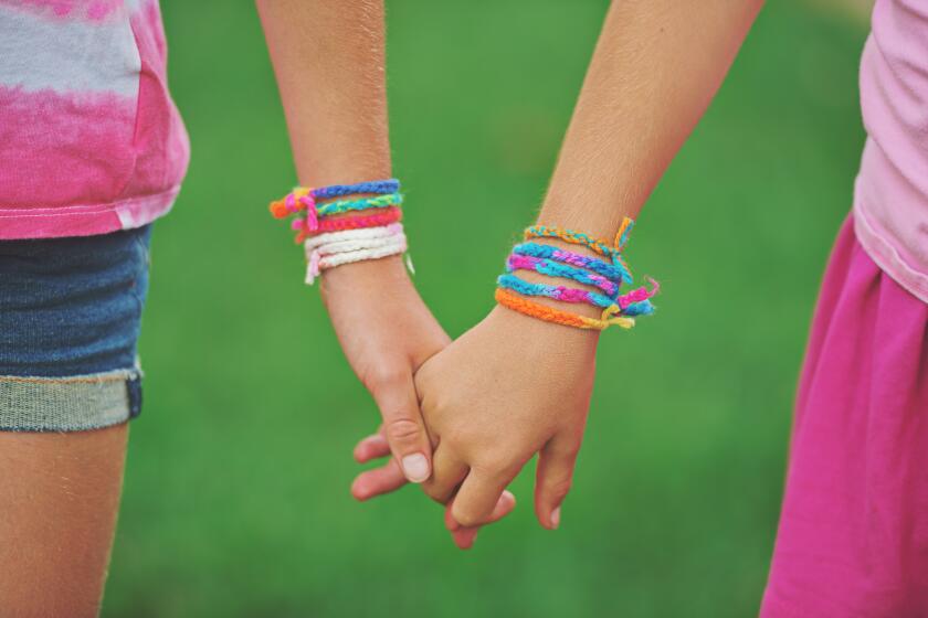 Two young sisters holding hands wearing friendship bracelets they made themselves using colorful yarn.