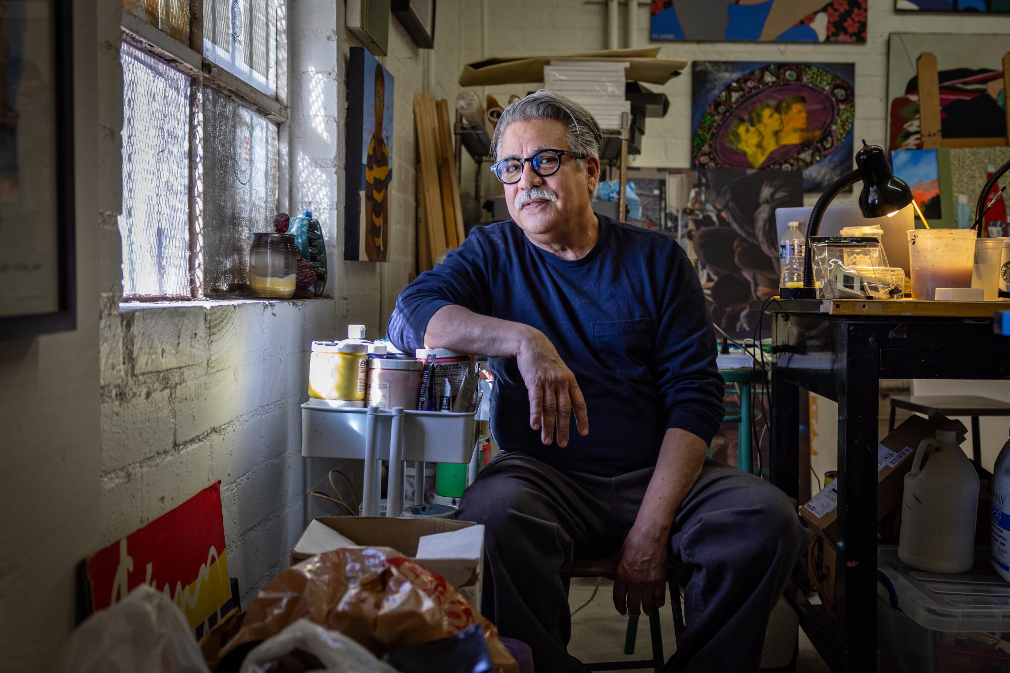 Joey Terrill, a Chicano man in his late 60s, sits amid containers of paint and colorful collages in a cluttered studio.