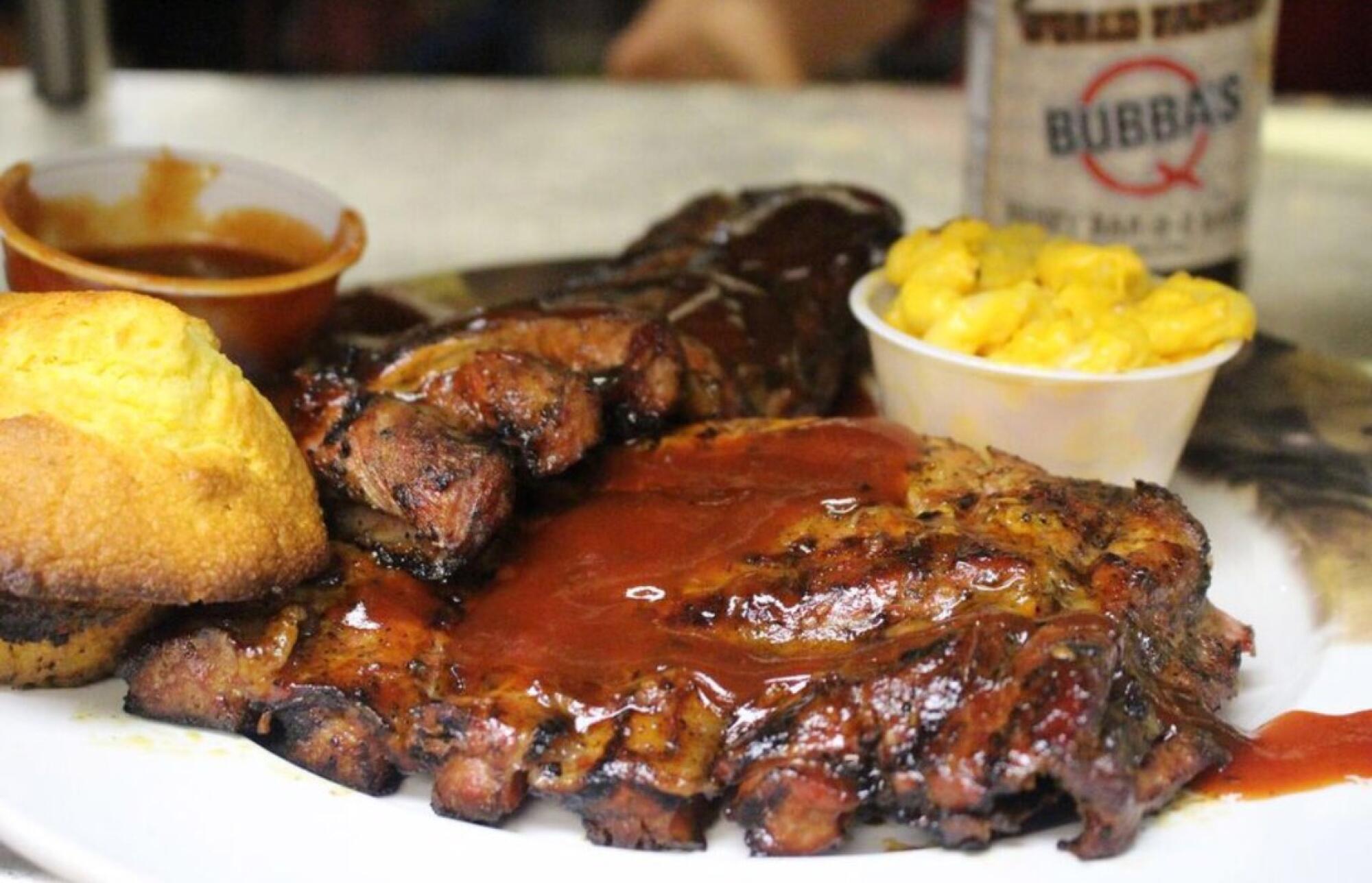A plate of ribs with bread, mac and cheese and a cup of extra sauce on the side